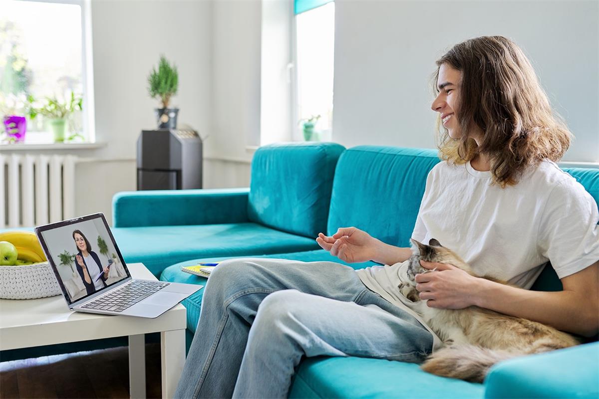 A young man attends a telehealth mental health appointment while petting his emotional support cat beside him on the couch.